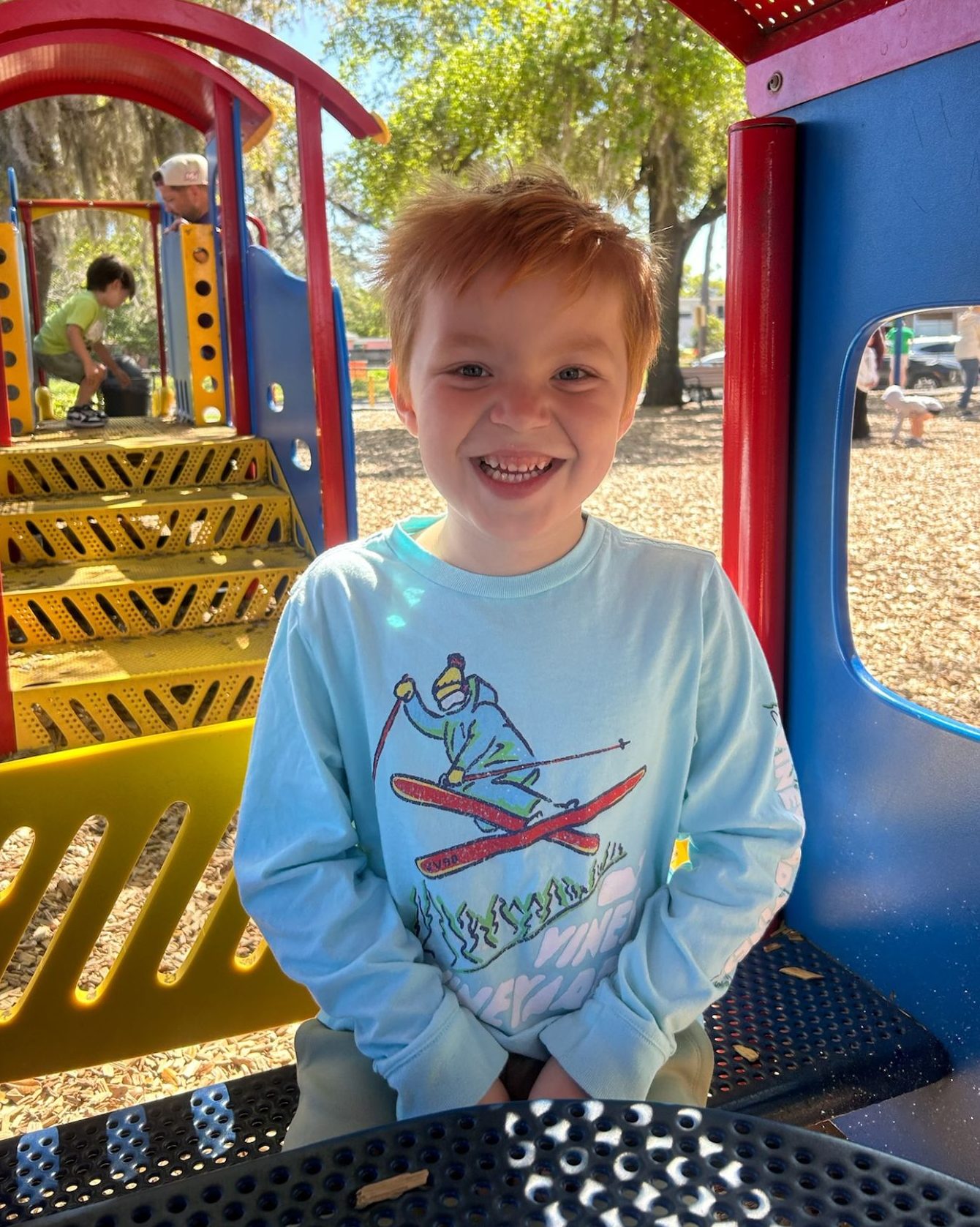 A young child leans joyfully toward the camera at a playground, smiling brightly.