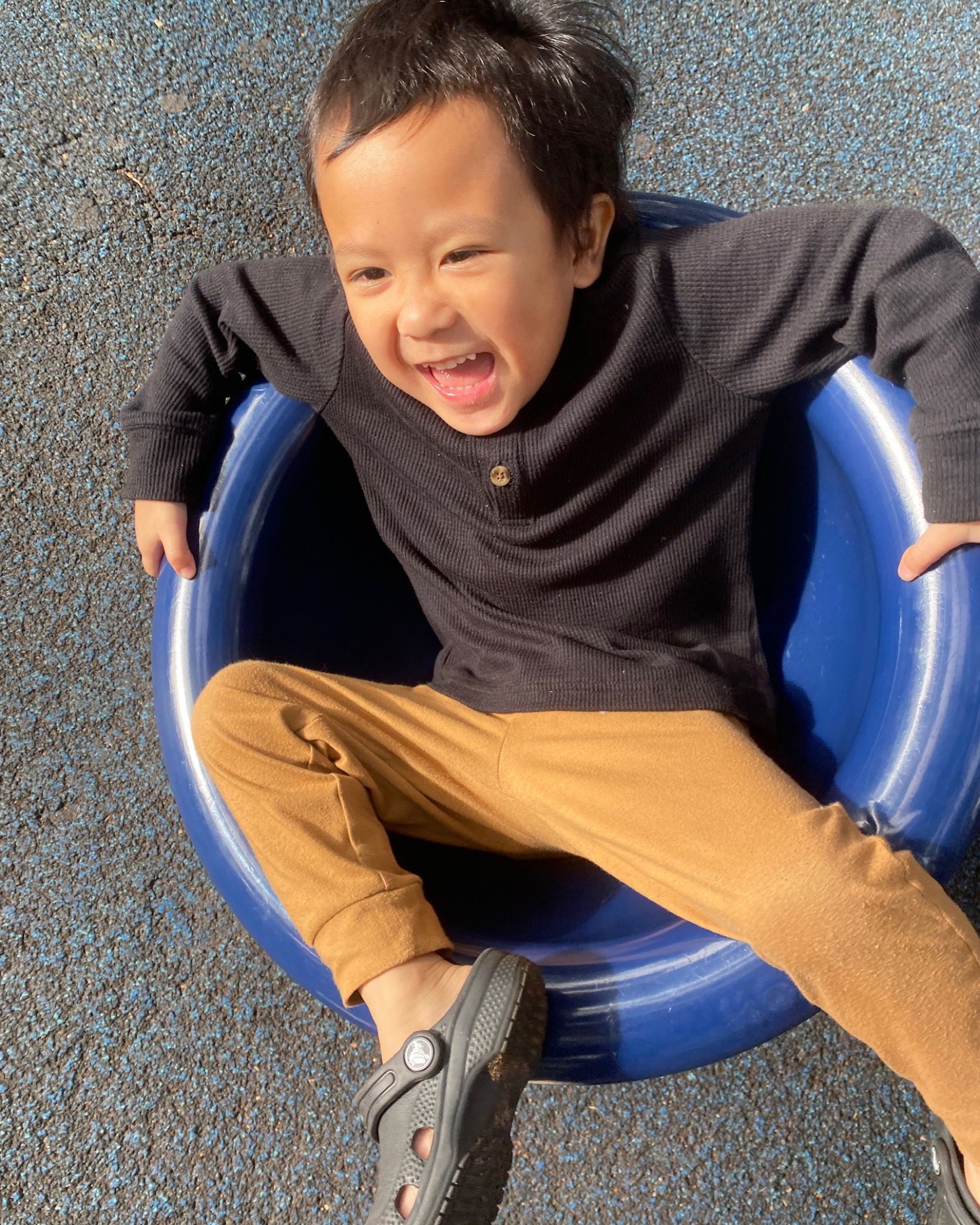 A laughing toddler reclines inside a blue spinning play seat on a playground.