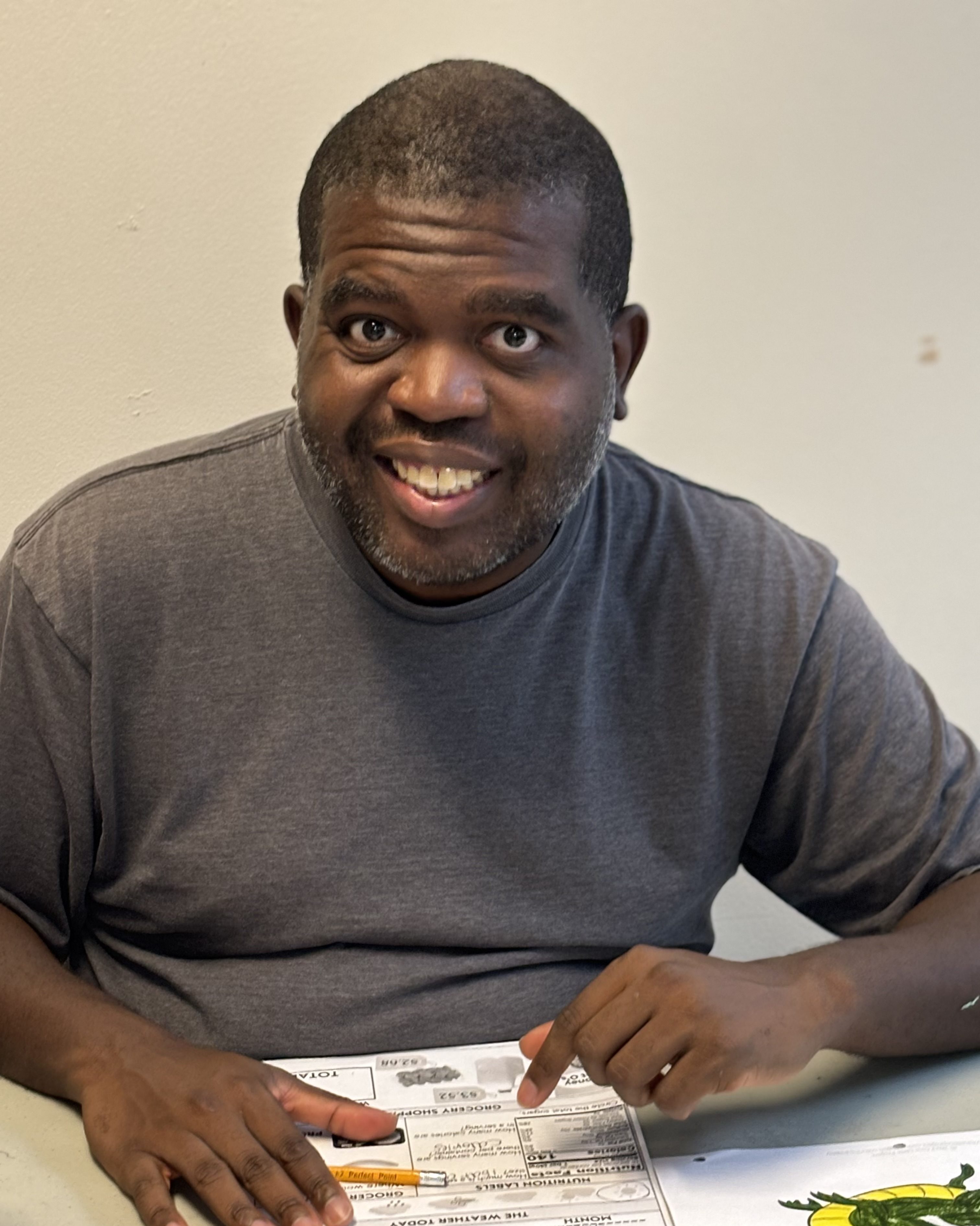 A man smiles at the camera. He is sitting at a table with a worksheet and pencil in front of him.