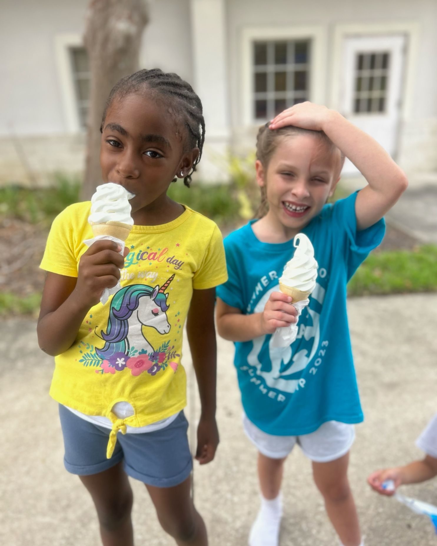 Two children stand together eating ice cream. One is holding the ice cream to her mouth and the other is smiling and laughing as she poses with the ice cream in front of her.